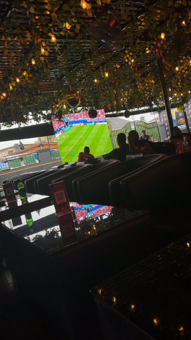 A dimly lit bar or sports venue showing a football match projected on a large screen, with people sitting at tables and decorative foliage hanging from the ceiling.