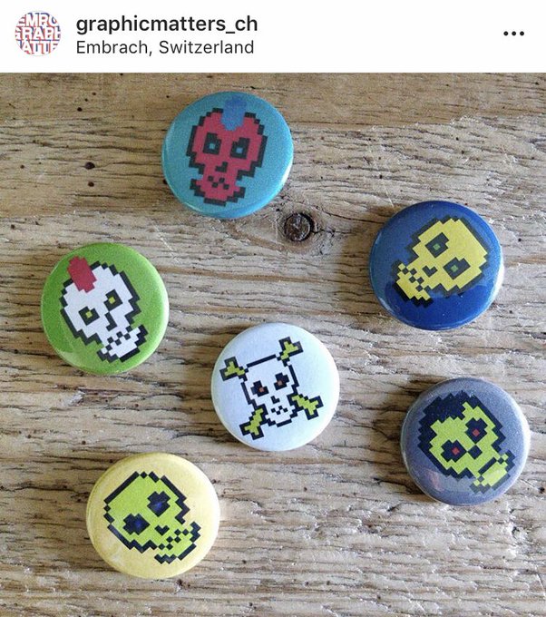 A collection of six pixel-art skull pin badges in various colors (red, green, yellow, white, blue, gray) arranged on a wooden surface, posted by Instagram user 'graphicmatters_ch' from Embrach, Switzerland.