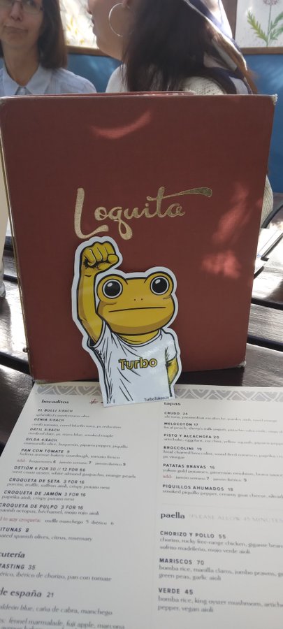 A red menu for "Loquita" Spanish restaurant rests on a table, with a yellow cartoon frog sticker wearing a shirt that says "Turbo" placed on top.