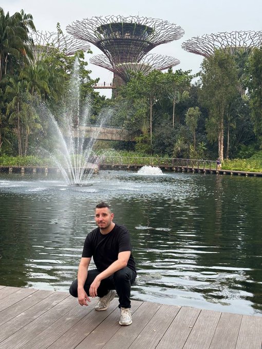 A man in a black t-shirt squats on a wooden deck in front of a pond with fountains, with the iconic Supertree Grove structures of Gardens by the Bay in Singapore visible in the background.