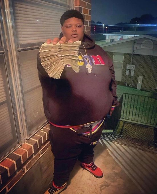 A young man stands outside at night on a porch, holding up a thick fan of cash toward the camera while wearing a dark hoodie and red sneakers.