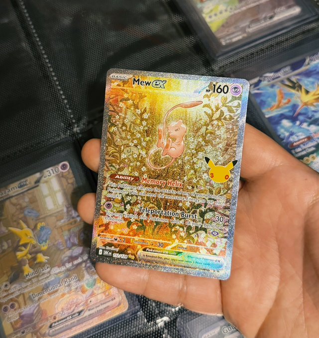 A hand holds a holographic Pokémon trading card featuring 'Mew ex' with 160 HP, displaying abilities 'Memory Walk' and 'Teleportation Burst' alongside other cards in a binder.