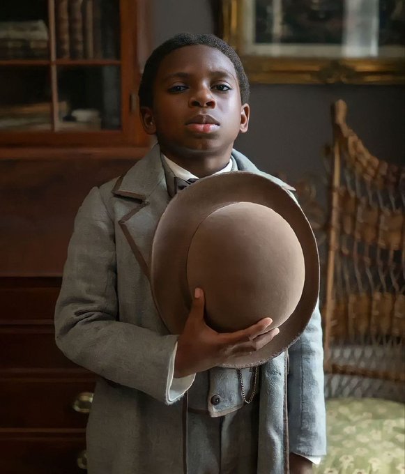 A young boy dressed in a vintage gray suit holds a brown felt hat in his hand, standing in a classic room with wooden furniture and a wicker chair.