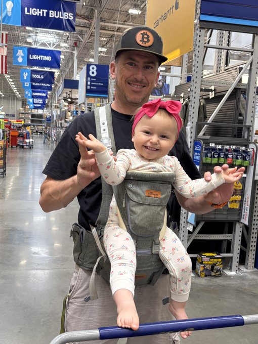 A smiling man wearing a Bitcoin cap carries a baby in a pink headband in a baby carrier while shopping at a hardware store in the Light Bulbs section.