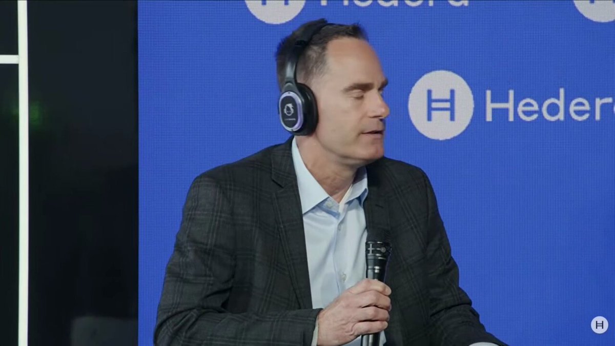 The image features a man wearing headphones while sitting at a desk or podium. He appears to be speaking into a microphone, possibly giving a presentation or participating in an interview. The background of the image shows a blue wall with the words "hedera" written on it, indicating that this is likely taking place during a meeting or event related to Hedera's digital transformation efforts.