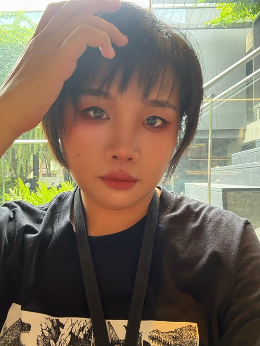 A young woman with dark hair taking a selfie, resting her hand on her head, wearing a black graphic t-shirt with a lanyard, with a modern building and greenery visible in the background.