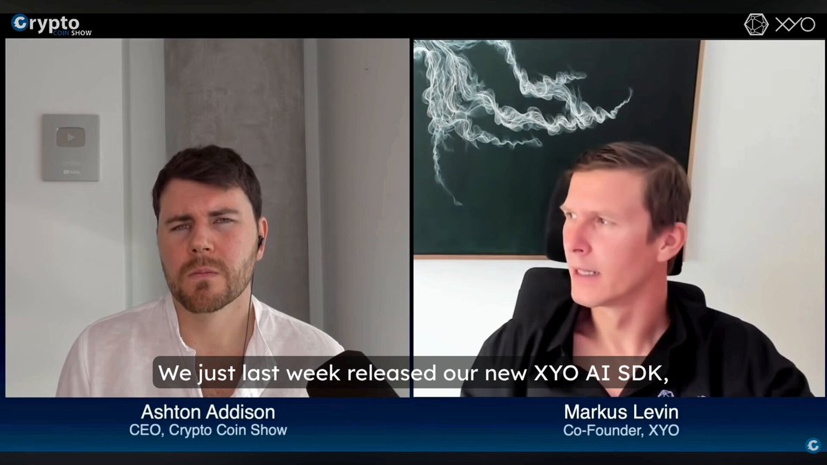 The image shows a screen with two men sitting across from each other, likely having a conversation or participating in an interview. The man on the left side of the frame has his arms crossed, while the man on the right side leans forward attentively. Both individuals are wearing white shirts and appear to be engaged in a discussion about the new XYO AI SDK.