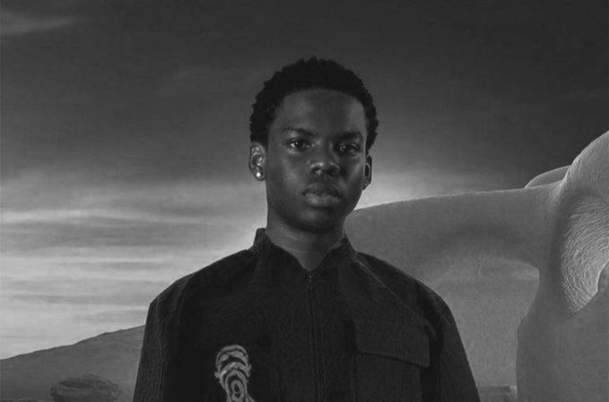 A black-and-white portrait of a young man in a dark zip-up jacket standing against a dramatic cloudy sky with a large, curved stone structure behind him.