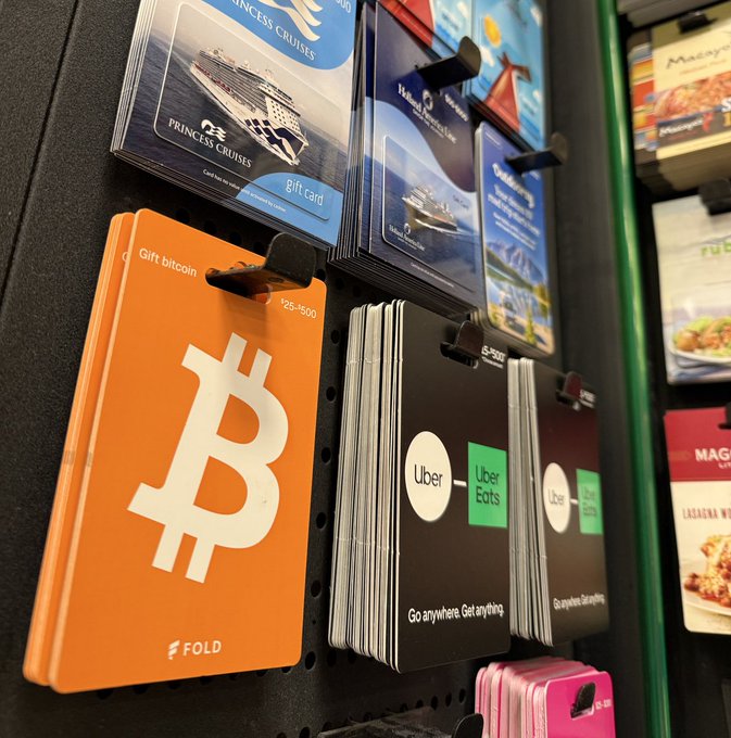A store display shows branded gift cards including a prominent orange "Gift bitcoin" card by Fold with the Bitcoin logo, alongside other cards from Princess Cruises, Uber Eats, and Maggiano's. The image visually supports the claim about Bitcoin gift cards being sold at retail locations across the U.S., demonstrating mainstream adoption of cryptocurrency as a payment option.