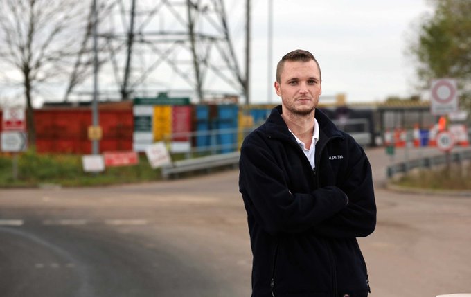 A man with short hair wearing a black fleece jacket stands with arms crossed in front of a construction site with electrical transmission towers and safety signage.