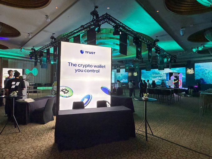 An event booth displaying a lit 'TRUST' banner reading 'The crypto wallet you control,' set in a large hall with stage lighting and other attendees visible in the background.