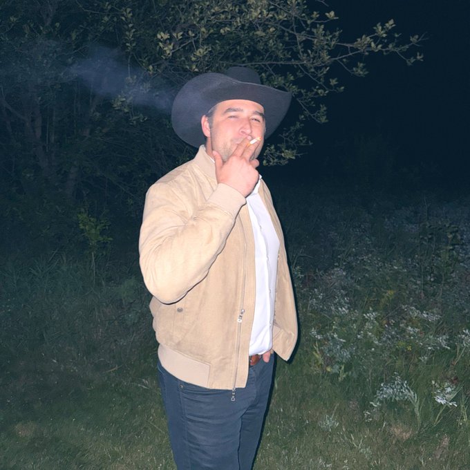 A man wearing a black cowboy hat and tan jacket smokes a cigarette outdoors at night, with smoke visible and a leafy background behind him.