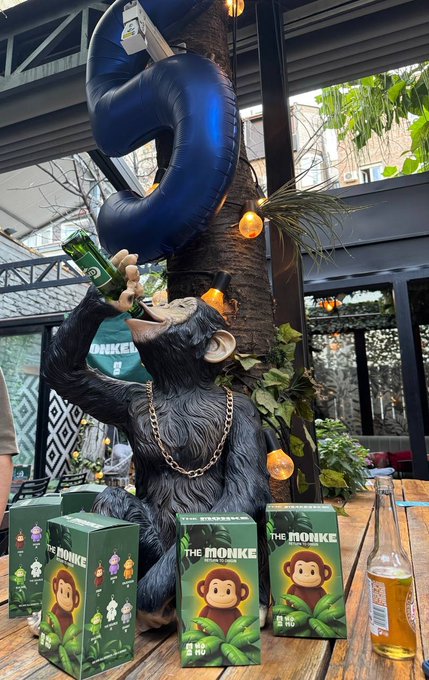 A black chimpanzee statue wearing a gold chain drinks from a green 'The Monke' beverage bottle while holding a large number '5' balloon, surrounded by green product boxes on a wooden table at an outdoor venue.
