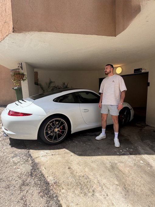 A man stands proudly next to a white Porsche 911 in a garage, casually dressed in a striped t-shirt and shorts, greeting his friends. The post "Gm friends" reflects his relaxed, friendly demeanor as he poses with his sleek sports car.