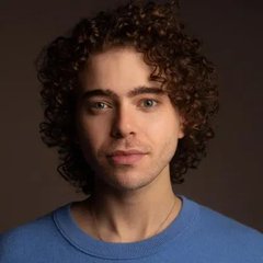 A portrait photograph of a young man with curly dark hair wearing a blue sweater against a brown background.