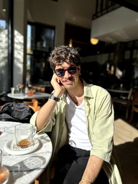 A young man with curly hair and sunglasses smiles while seated at an outdoor cafe table with a glass of amber liquid.