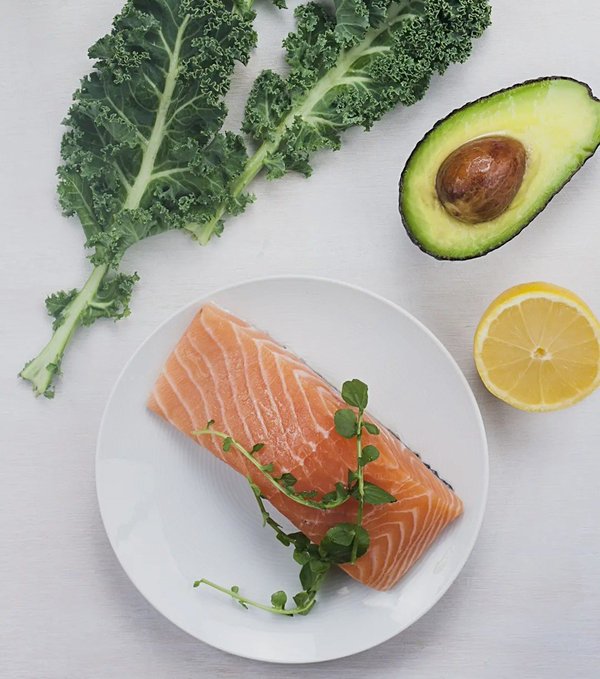 Overhead photograph of fresh ingredients including a raw salmon fillet on a plate garnished with herbs, kale leaves, a halved avocado, and a lemon slice.