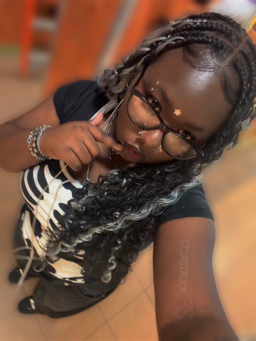 A woman with long braided hair, glasses, and star face decals poses for a selfie while wearing a black and white outfit and silver jewelry, with the watermark "SI-345TRUCK" visible on her arm. The caption playfully suggests a return to findom, asking if followers missed her and addressing "wallets and piggies," fitting the confident, commanding demeanor shown in the photo.