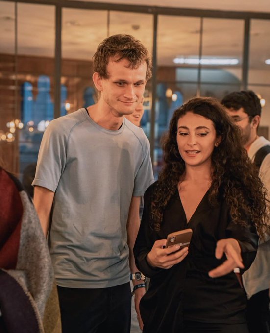 A man in a light blue t-shirt and a woman with long curly dark hair holding a smartphone stand together at an indoor social event.