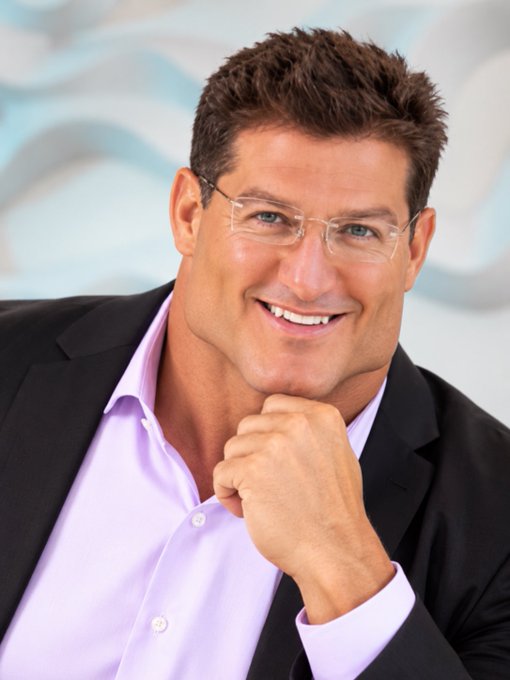 A professional headshot shows a smiling man with short brown hair and rimless glasses, wearing a light purple shirt and a black suit jacket against a light blue background.