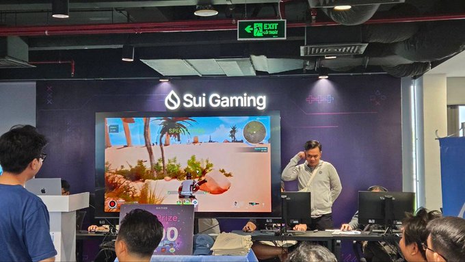 A gaming event booth for Sui Gaming featuring a large screen displaying a desert-themed video game, with attendees and a booth operator wearing a lanyard.
