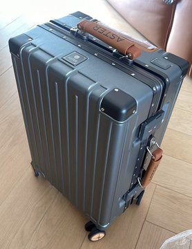 A sleek dark gray hardshell suitcase with a brown leather "ASTER" branded handle strap sits on a wooden floor, presented as a luxury gift package from Aster DEX. The premium luggage serves as a branded promotional item, likely intended for a giveaway or VIP recipient in the crypto community.
