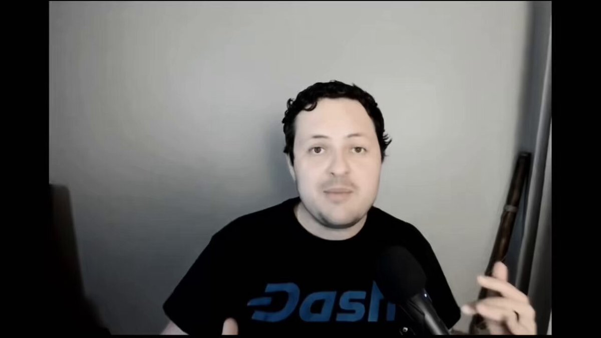 The image features a man wearing a black t-shirt with the word "Dash" written on it, holding a microphone and standing against a gray wall. The background of the image shows a computer monitor or screen behind him, suggesting that he might be in front of a TV or a computer setup. The overall atmosphere seems to be casual and relaxed, as if the man is engaging with an audience through a live stream or video call.