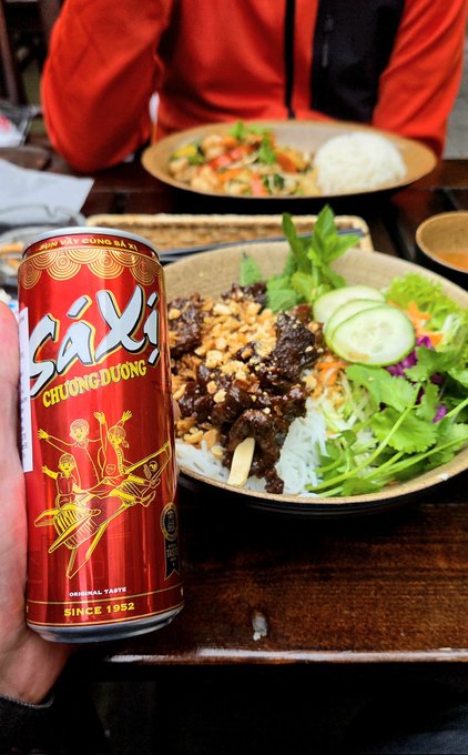 The image shows a person holding a can of SAXI Chonbokulonic soda, which is red with gold lettering. The person is sitting at a dining table with a bowl of food containing noodles, vegetables, and meat. There are also other bowls on the table, one in front of them and another behind it. A chair is visible near the table as well. The post about SALE has been mentioned, suggesting that someone might have posted this image to share their experience or recommend the soda.