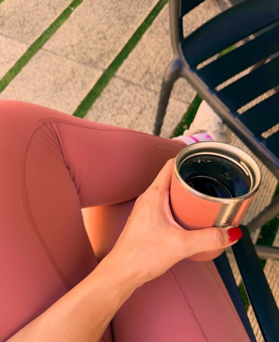 A first-person view of a person in pink athletic leggings holding a peach-colored tumbler with a dark beverage while sitting outdoors on a patio with dark chairs.