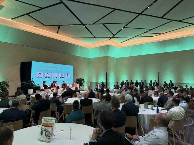 A photograph of a large conference or seminar room where attendees sit at round tables facing a panel and a presentation screen displaying headshots with text mentioning 'Getting investors to take the plunge into...'.
