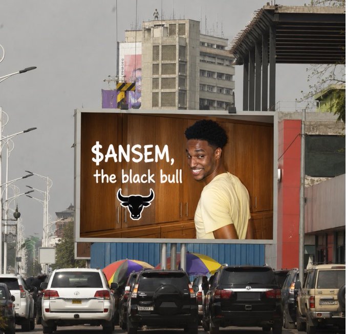 A large billboard stands prominently in a busy city street, featuring a smiling young Black man in a yellow t-shirt alongside the text "$ANSEM, the black bull" and a bull logo. The surrounding scene shows tall buildings, traffic with various cars, and colorful umbrellas below, illustrating the rented advertisement meant to catch the attention of @blknoiz06 as described in the post.