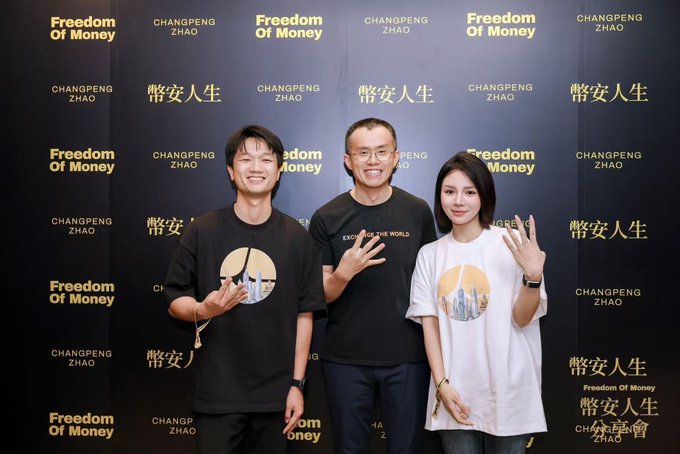 The image shows three people posing in front of a backdrop featuring the text "Freedom Of Money," "CHANGPENG ZHAO," and "幣安人生" (Binance Life). The man in the center, presumably Changpeng Zhao (CZ), the founder of Binance, is making a hand gesture alongside a young man on the left and a young woman on the right, both wearing t-shirts with Binance-themed designs featuring city skylines. This scene likely relates to the post about CZ's interaction with someone from the Aster project, showcasing his engagement with builders in the BNB Chain community.
