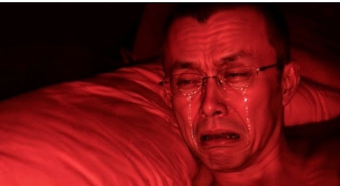 A man wearing glasses lies in bed bathed in red lighting, crying with visible tears streaming down his face.