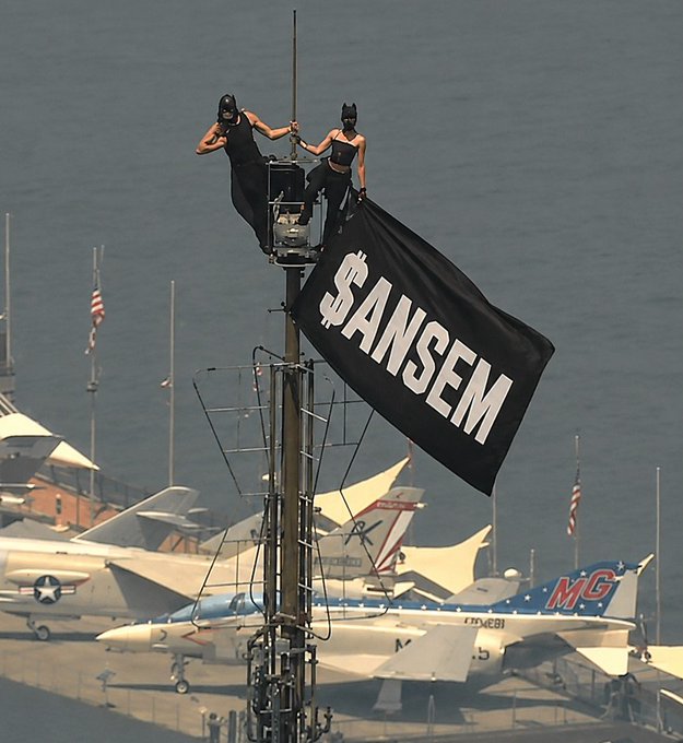 Two figures in black outfits and masks perched atop a mast, holding a large black flag reading '$ANSEM' at a marina with military aircraft displayed below.