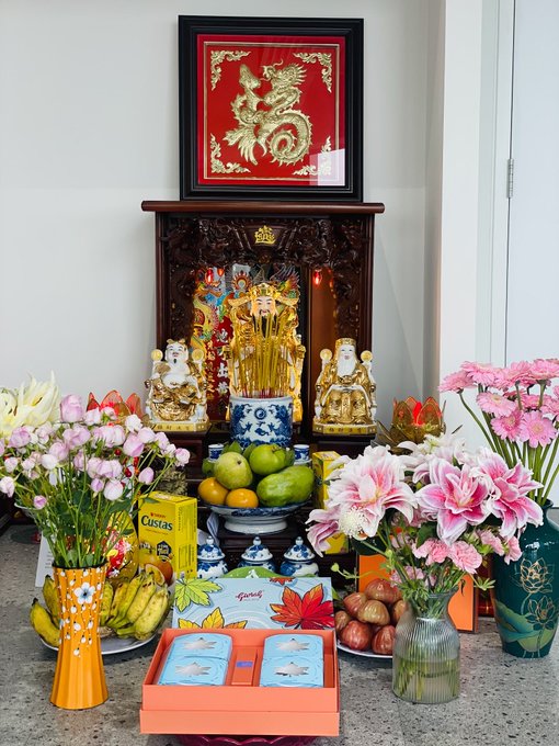 A traditional Chinese/Vietnamese ancestral altar features a golden dragon artwork, deity statues, burning incense, and offerings of fruits, flowers, and snacks including 'Custas' crackers.
