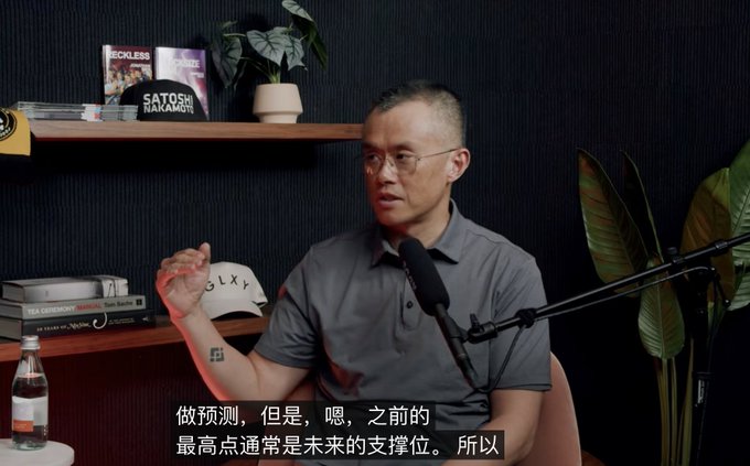 The image shows a man wearing glasses sitting at a desk with a microphone, likely recording an interview or podcast. The background features a black wall and a plant, creating a professional atmosphere for the discussion. There are several books scattered around the room, suggesting that the man might be researching or discussing various topics related to cryptocurrency, such as Bitcoin, Hyper Liquid, and AI.
