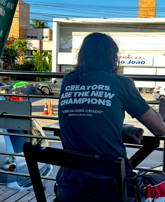 A person with long dark hair sits facing away from the camera wearing a navy shirt that reads "CREATORS ARE THE NEW CHAMPIONS" and "CRIE OU SERÁ CRIADO - CREATE CLUB EST.2025," overlooking a street with a "FARMÁCIAS São João" sign.