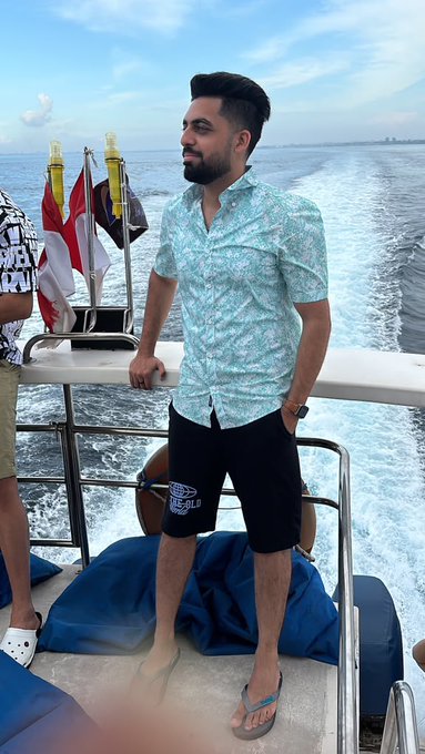 A man with a beard stands on a boat wearing a blue patterned shirt and dark shorts, looking out at the water with the boat's wake visible behind him.