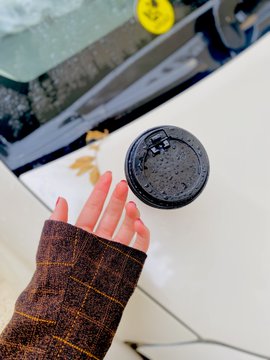 A hand wearing a plaid brown sleeve with red fingernails balances a black coffee cup lid on its fingertips, with a glass-topped table visible in the background.