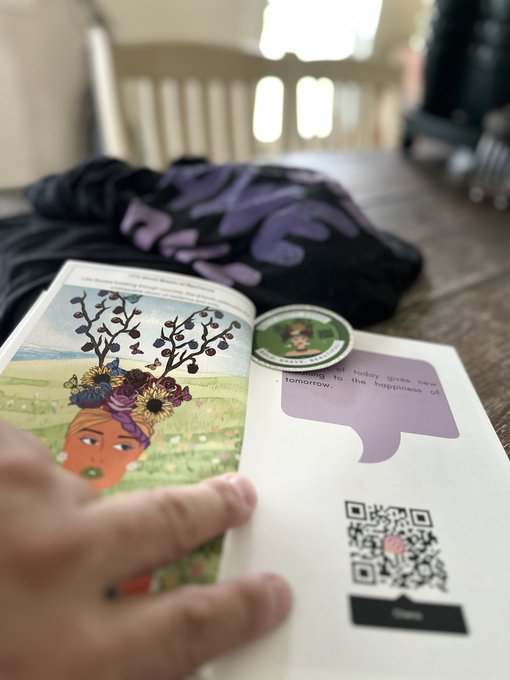 A person's hand turns the page of a children's book featuring an illustration of a woman with a flower crown and bird-filled trees, with a QR code displayed on the opposite page.