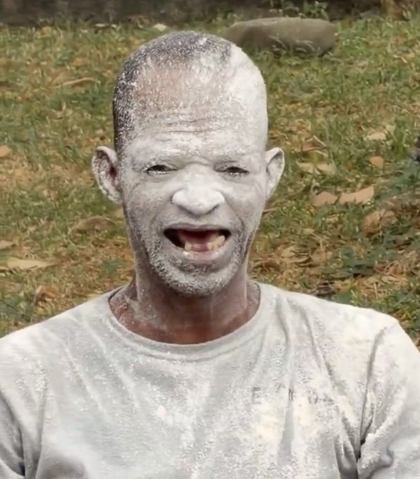 A close-up photo of a smiling man with his head and gray t-shirt completely covered in white powder or ash, standing outdoors on grass.