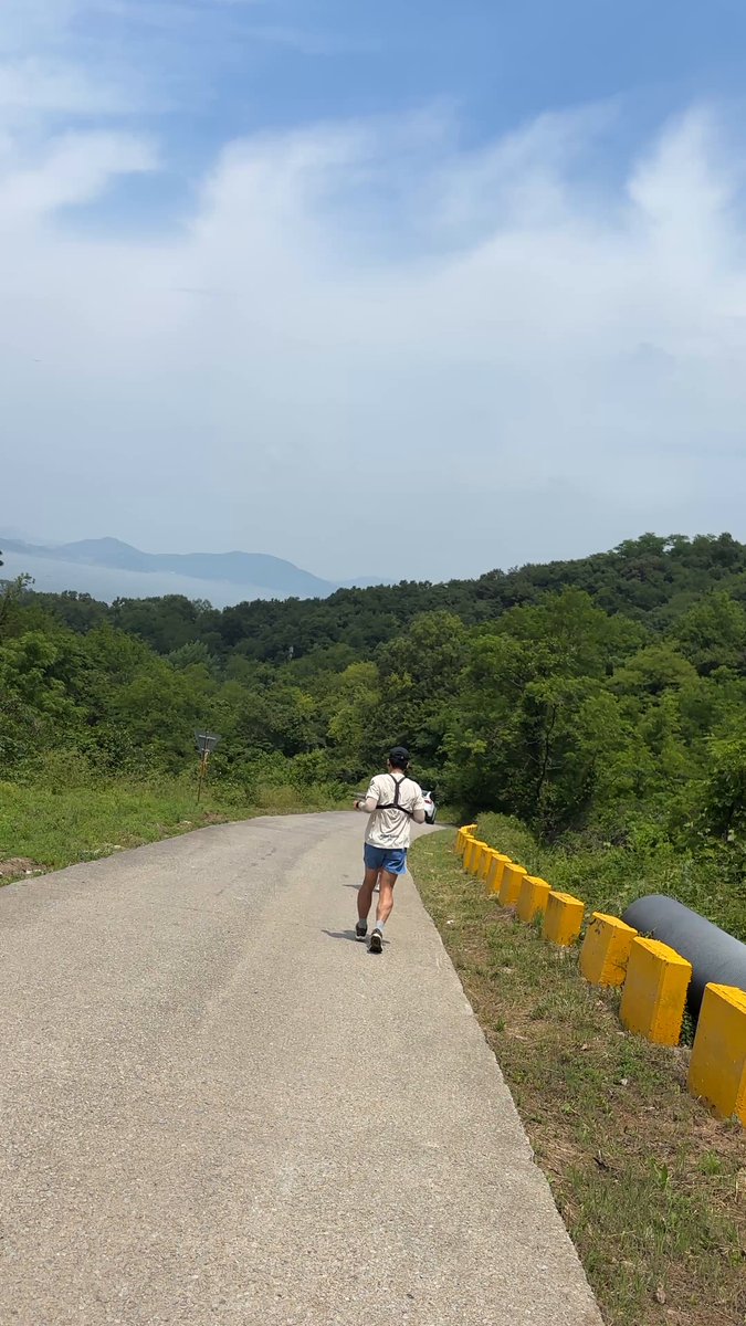 The image shows a person riding a scooter down a road, with their back facing the camera. The road appears to be made of concrete or asphalt, as there are no visible signs of grass or dirt. In the background, there are trees lining the side of the road, adding some greenery to the scene. The post accompanying this image is titled "1年ぶりの韓国！ 韓国STEPNerのJuneとホンジュさんと15kmランでついてきます". This post seems to be promoting a scooter ride or possibly an event related to scooters.