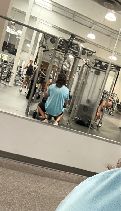 A selfie-style photo taken in a gym mirror showing a person sitting at a workout machine, with their own reflection visible in the foreground.