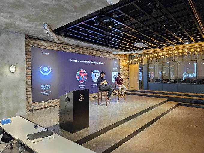 An indoor event space with a stage where two people are seated having a Fireside Chat with Varun Vadhwa, H3ro6h, displayed on a large screen with the message 'Where FX happens.'