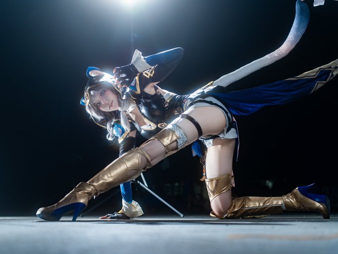 A cosplayer poses dramatically in an elaborate blue and gold costume with thigh-high boots, cat ears, and a long tail, capturing the feline grace of #Cipher from League of Legends' Star Guardian skin line.