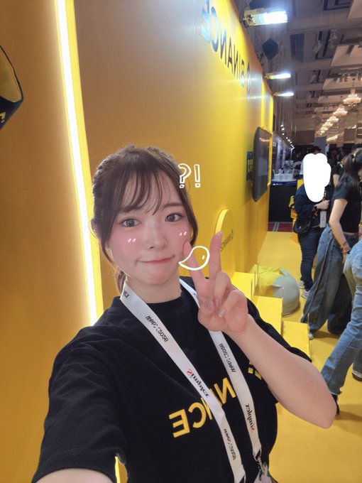 A selfie of a young woman at what appears to be a Binance or cryptocurrency convention, wearing a black shirt and lanyard, with a cute '? !' filter applied to her face.