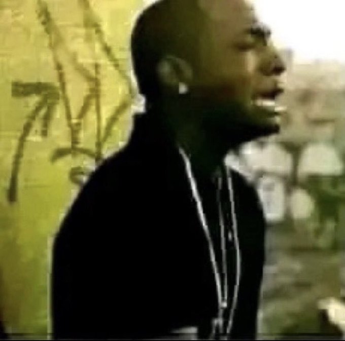 A low-resolution image of a man wearing a dark jacket and silver chains, appearing to rap or sing outdoors against a graffiti-covered wall.