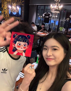 A young woman poses for a selfie in a restaurant while holding up a small painting of a cartoonish figure with dark hair, with chandeliers and other diners visible in the background.