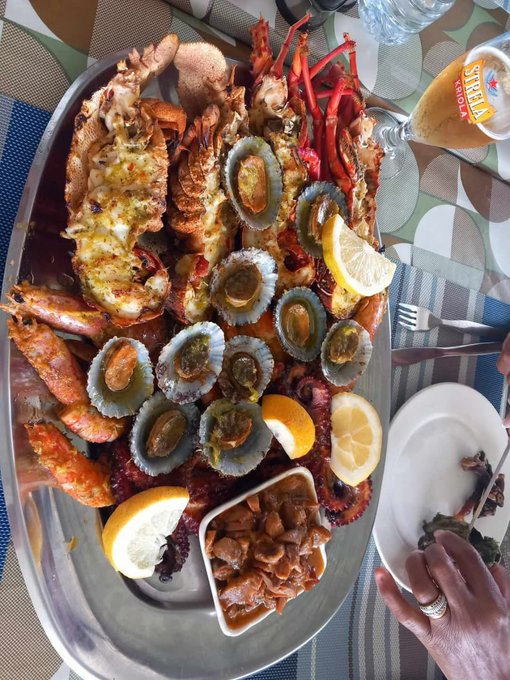 A large silver platter is filled with seafood, including lobster, crab, and abalone, accompanied by lemon wedges and a Stella Artois beer glass.