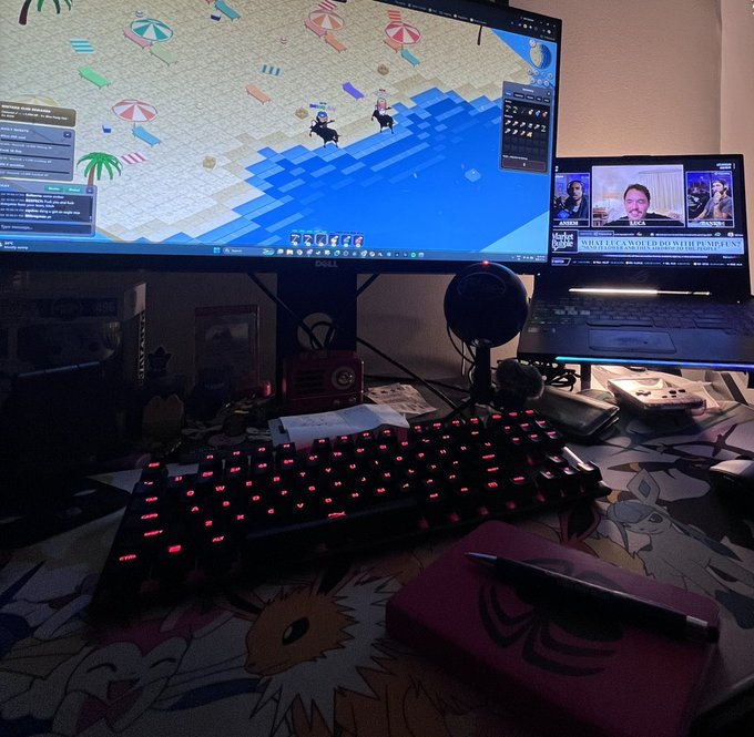 A dimly lit gaming setup with a glowing red backlit keyboard, a Dell monitor displaying a top-down pixel game on a beach, and a laptop showing a YouTube video.